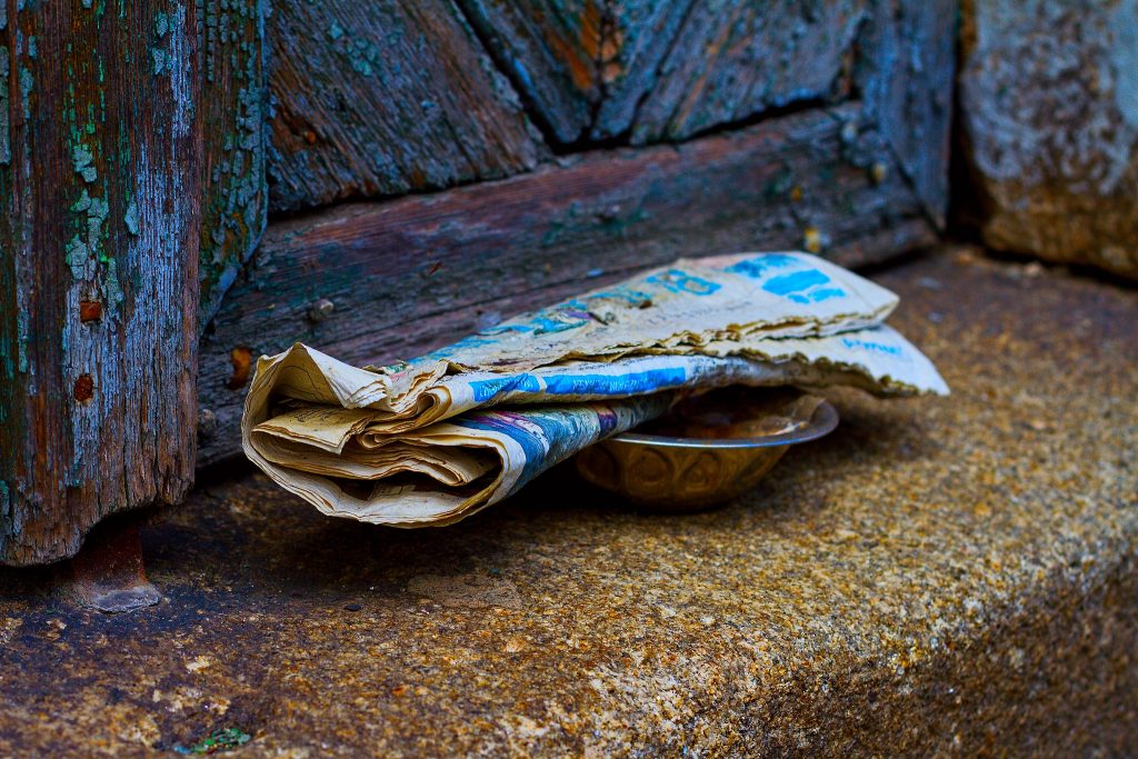 Eine seht alte Zeitung liegt in einer Schüssel aus Blech auf einem Treppenvorsprung. Die Farbe der ehemals blauen Tür blättert bereits ab.