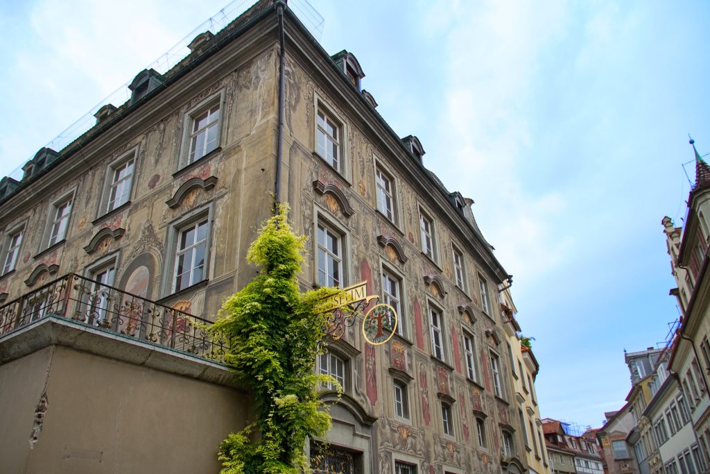 Ein altes, aber gut erhaltenes Haus in der Lindauer Innenstadt mit vielen Fenstern und Bemalung an der Außenseite des Gebäides. Blauer Himmel über dem Dach. Efeu rankt in der linken Bildhälfte im Vordergrund am Gebäude empor. Ein altes Schild mit der Aufschrift Museum ist im Efeu eingewachsen. Ein Balkon, an dem das Efeu ebenfalls emporwächst, ist in der linken unteren Bildhälfte zu sehen.