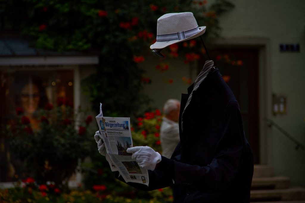 Ein Mann in der Innenstadt, der ohne Kopf eine Zeitung liest. Der fehlende Kopf ist damit erklärt, dass dieser in dem übergroßen Jackett steckt. Dadurch entsteht der "Kopflos"-Effekt. Im Hintegrund ist ein weiterer Mann zu sehen sowie die Gebäude der Lindauer Innenstadt.